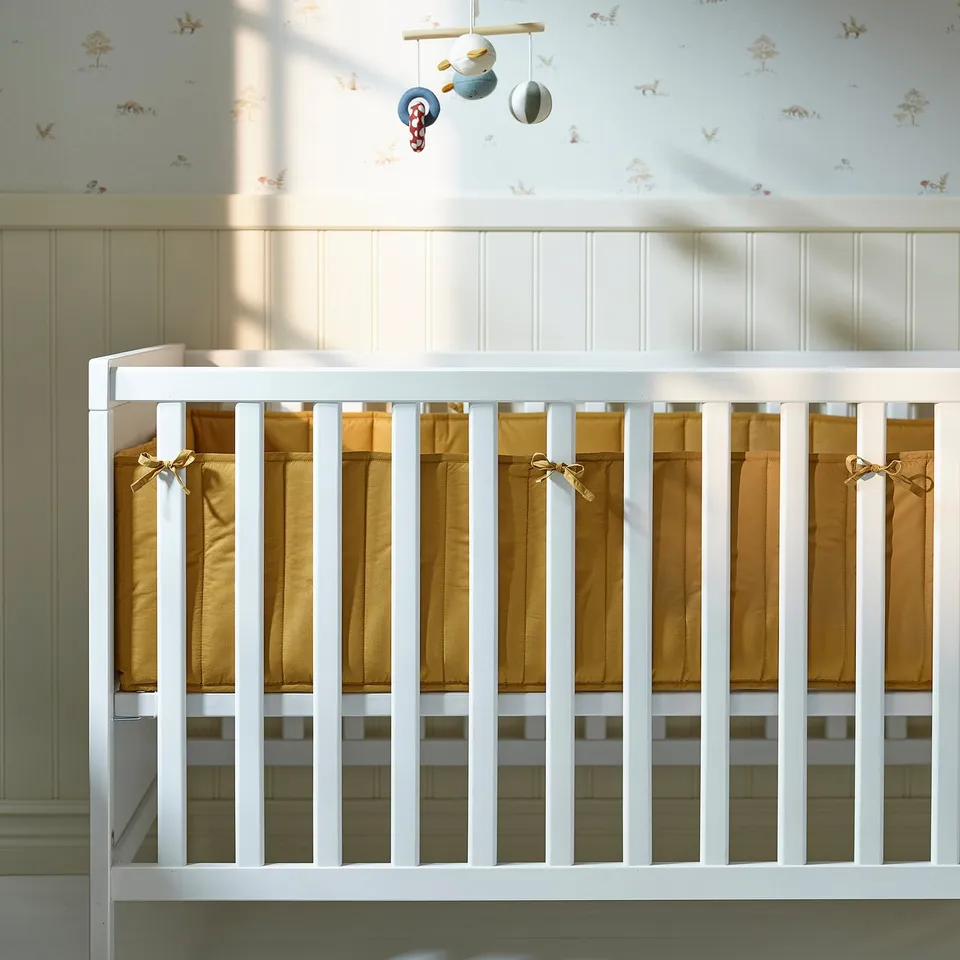 White crib with beige pads & decorative ribbons, mobile above.