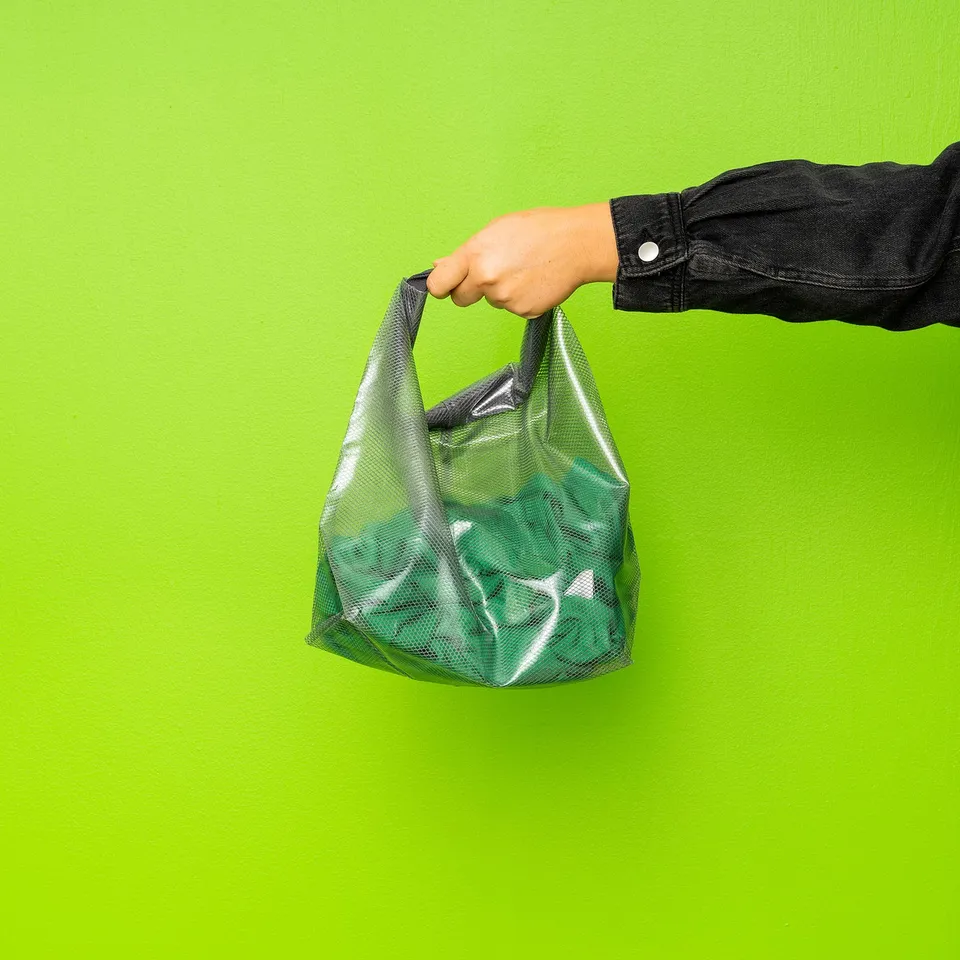 Hand holding green, mesh, drawstring bag against green wall.