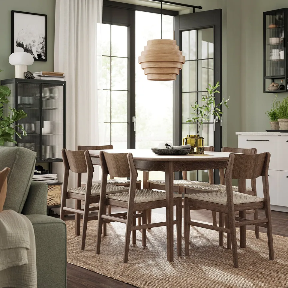 Modern dining area with SKANSNÄS wooden table, six matching chairs, and green walls. Large window with curtains, pendant light, and plants.