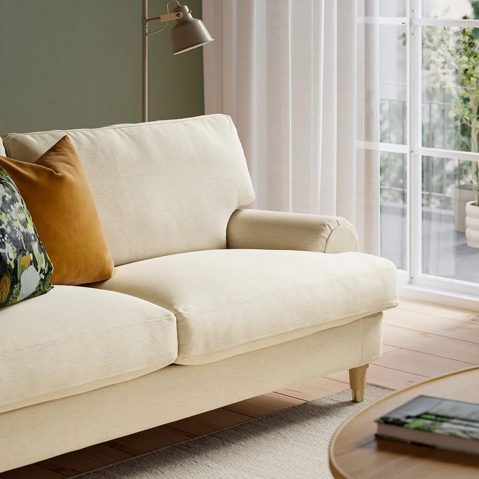 Beige sofa with brass feet, light wood floor, curtained window. Soft cushions, two decorative pillows.