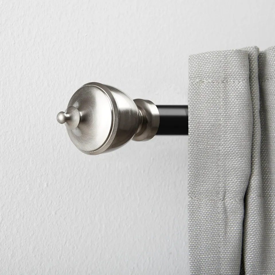A silver metallic finial firmly attached to a curtain rod, holding light grey curtains against a white wall.