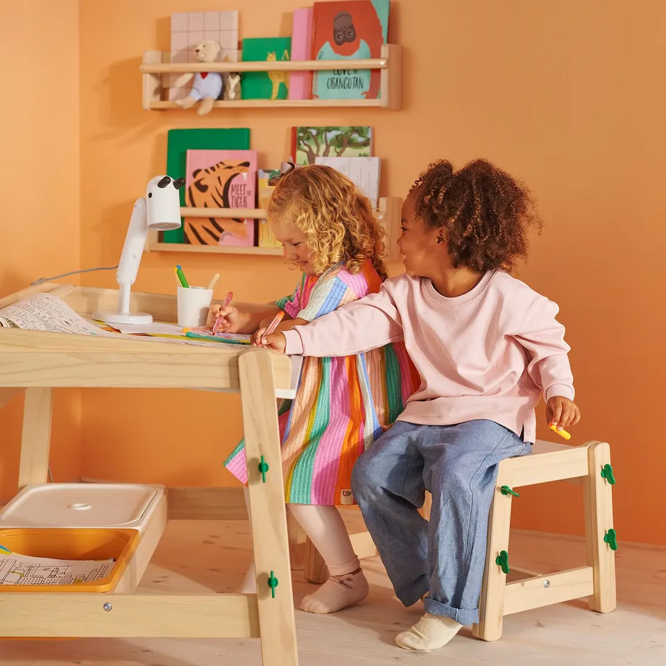 Two kids sitting at a height-adjustable FLISAT bench, drawing and using a desk, with colourful books and toys on shelves.