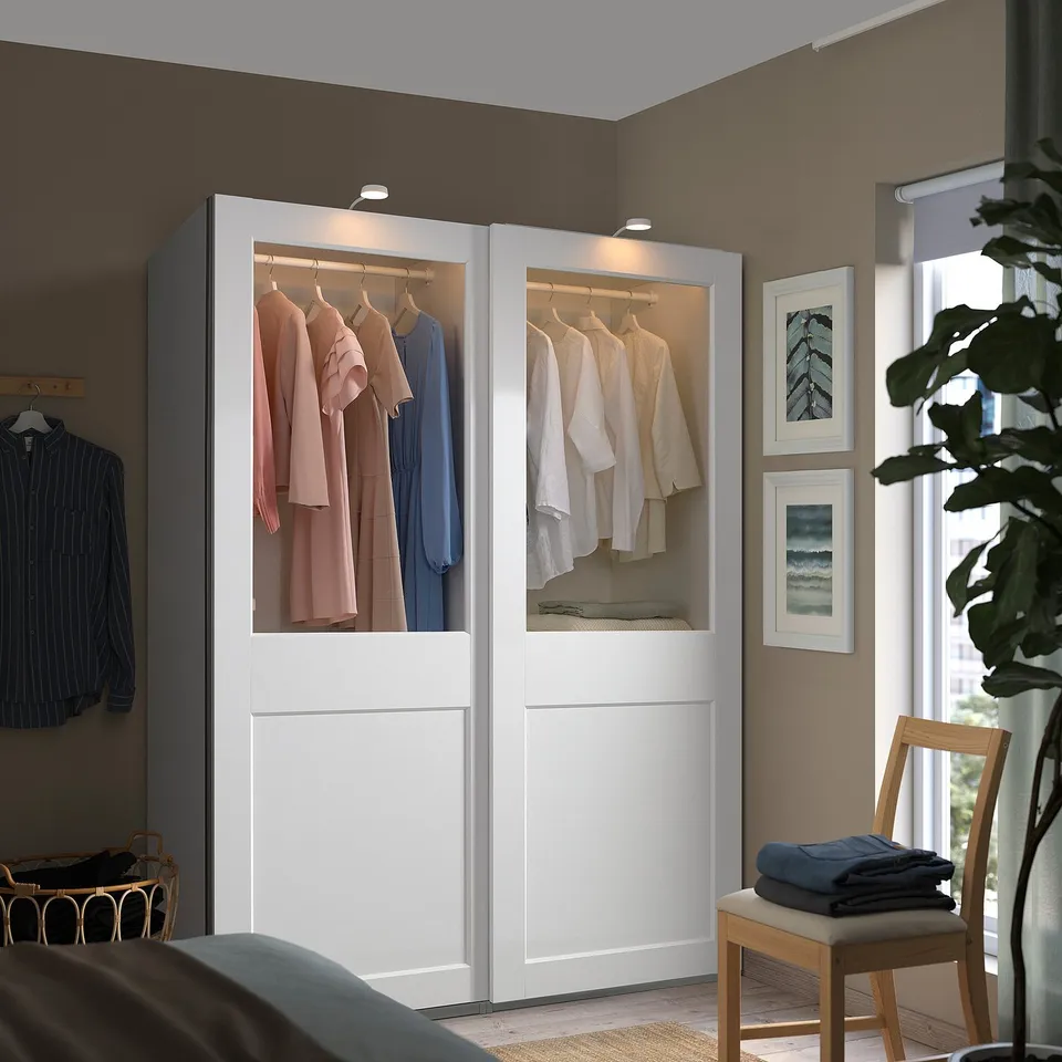 White PAX wardrobe with glass doors, light clothes, blue shirt on rack, dark clothes on bench, plant, two pictures.