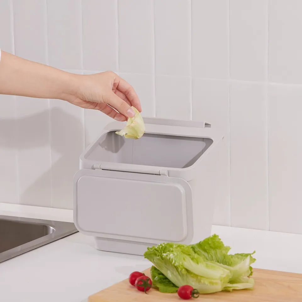 Hand placing food waste into a white SKOLÄST bin on a countertop, next to fresh vegetables.