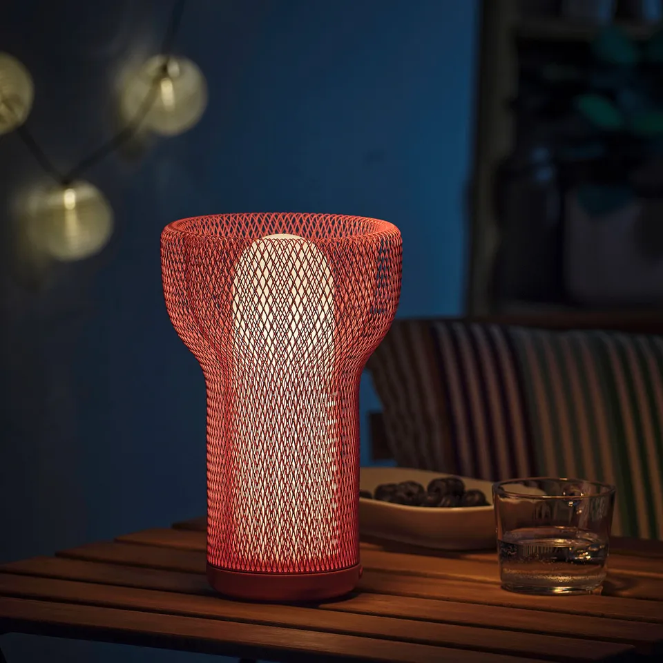 Unique red mesh lamp on table with lights, glass, and cushions. Light casts warm glow, ideal for indoor/outdoor use.