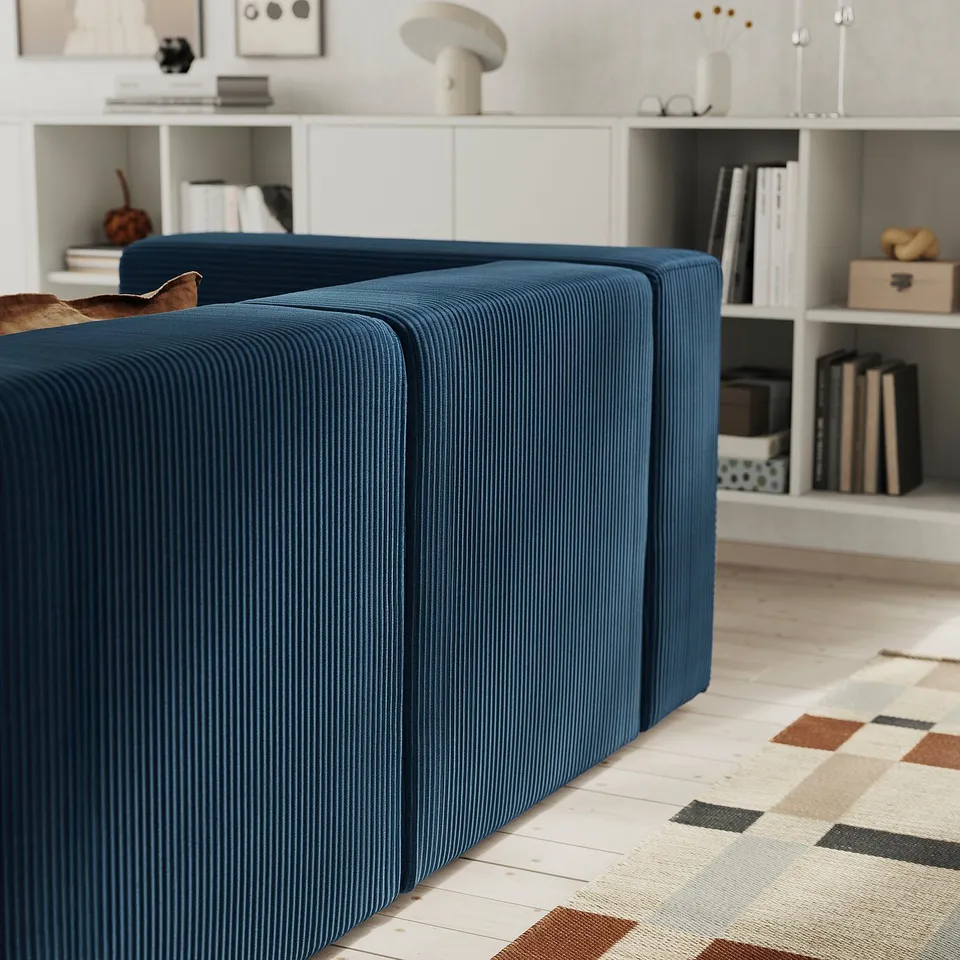 Modern living room with dark blue, corduroy modular sofa against a wall, white bookshelf with various items.