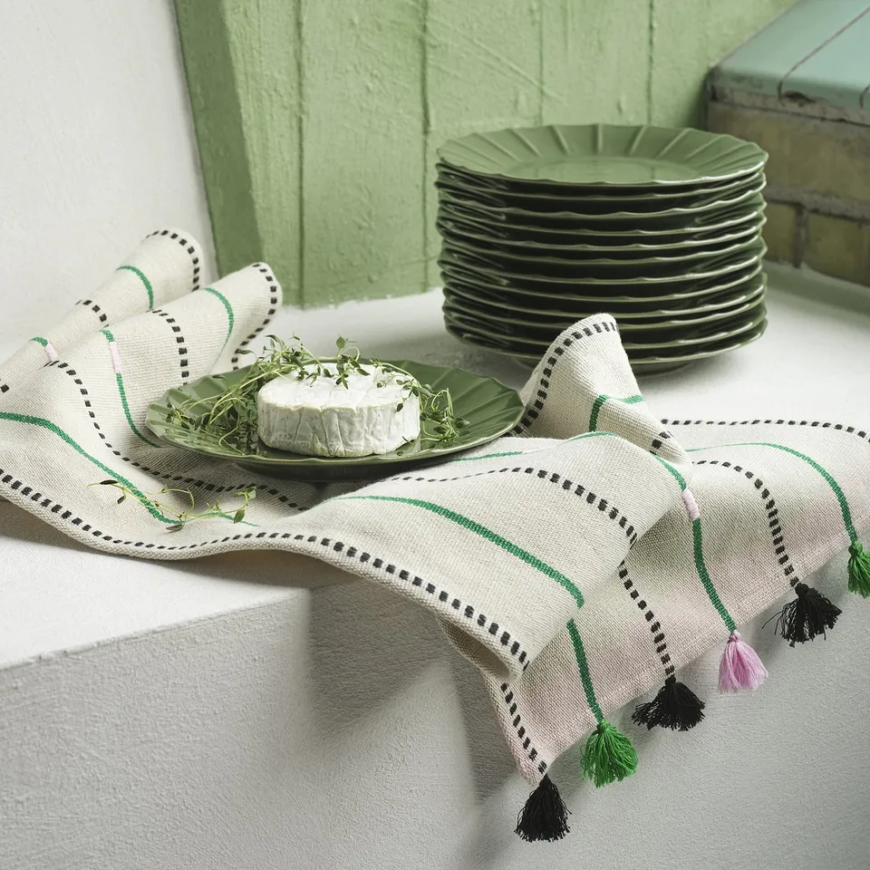 A green stoneware plate with embossed edge on a white cloth with stripes and tassels. Behind it, a stack of similar plates.