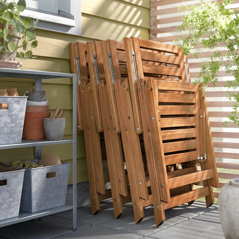 Stacked brown wood NÄMMARÖ chairs, folded, outdoors against green wall.