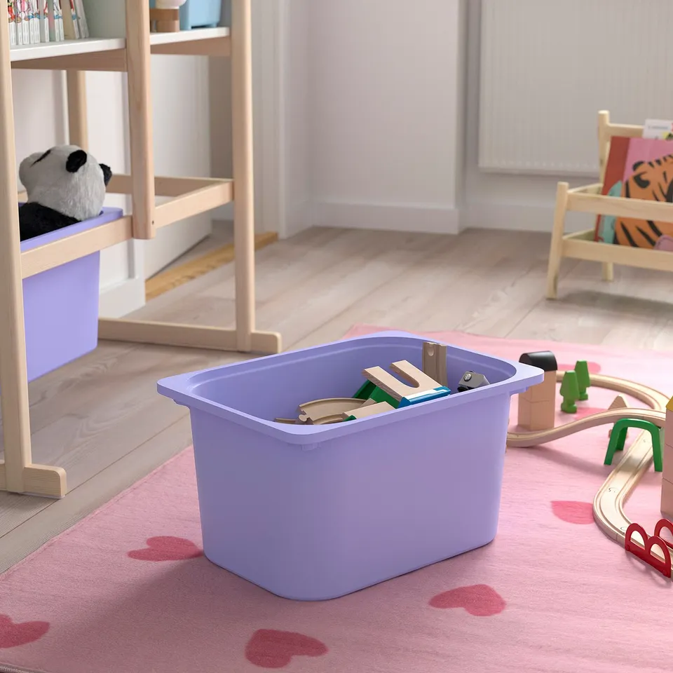 Purple bin with toys on pink rug, books, and stuffed animal behind, partial train set right.