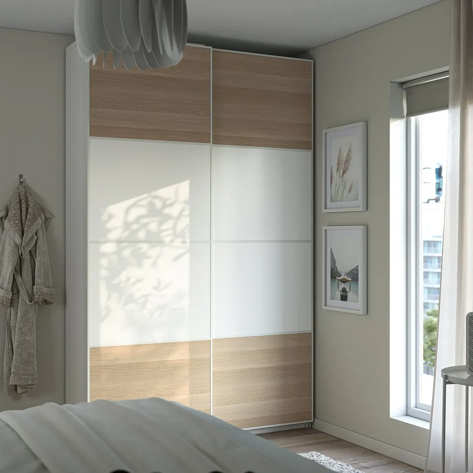 Modern bedroom with white wooden PAX wardrobe, grey robe, and light-colored bedding.