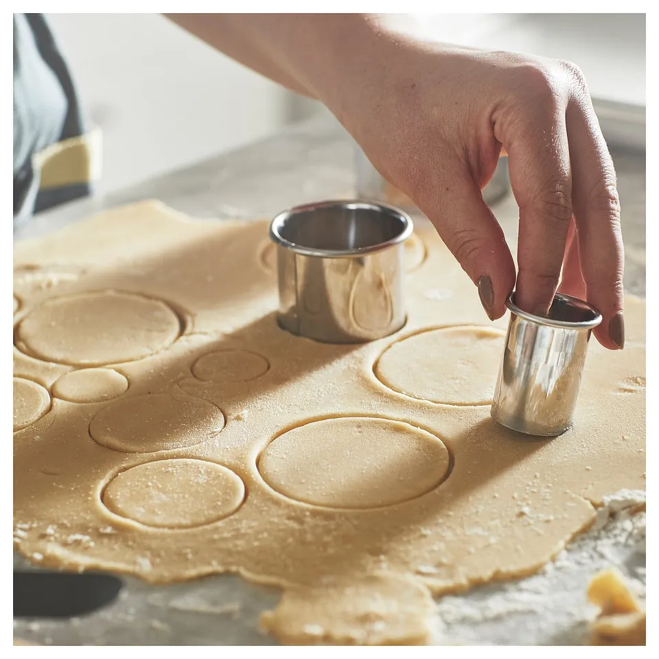 Silver circular cookie cutter used on rolled dough. Hand uses metal cutter to cut shapes from light-brown dough.
