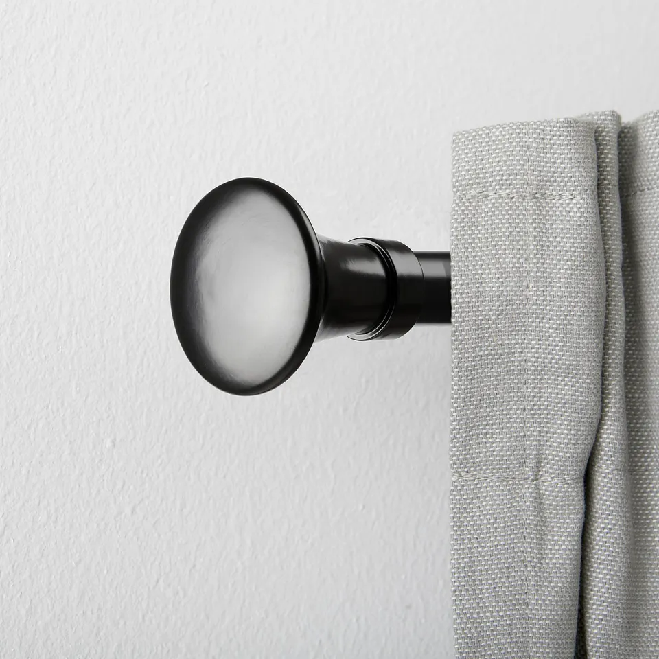 Black curtain rod finial with trumpet shape, holding light grey curtain against white wall.