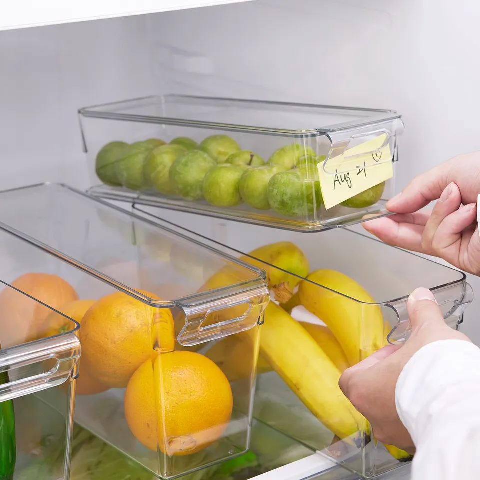 Clear fridge storage boxes organise fruits and keep track of storage dates. A hand holds the box with labelled limes.