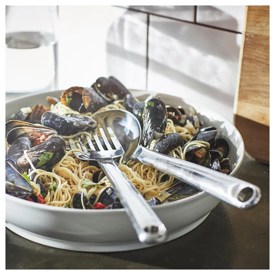 A white bowl of cooked spaghetti and mussels with stainless steel FINMAT fork and spoon, featuring curved handles for easy use.