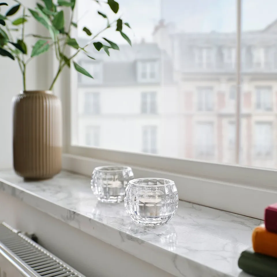 Two clear FRÖJDA glass candle holders on a marble windowsill, smaller than a vase with greenery.