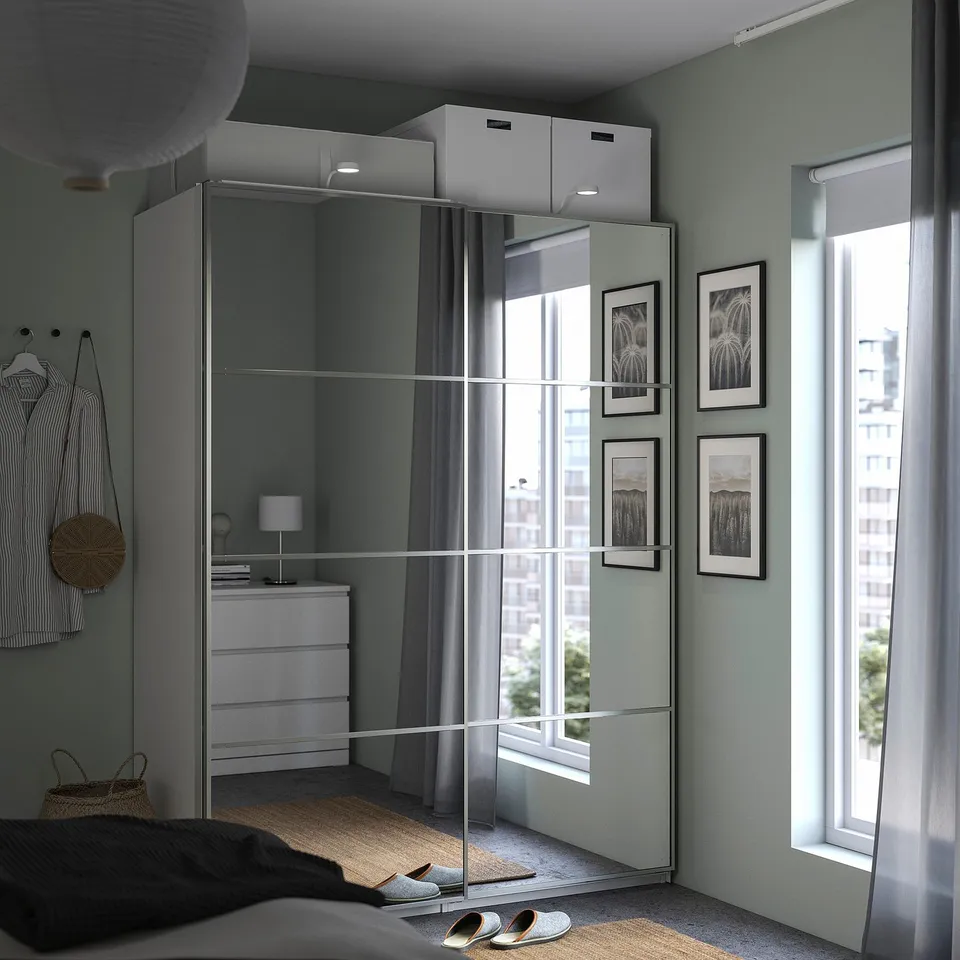 Modern bedroom featuring PAX wardrobe with mirrored sliding doors, grey fabric chest next to it, and white shelves with boxed storage above.