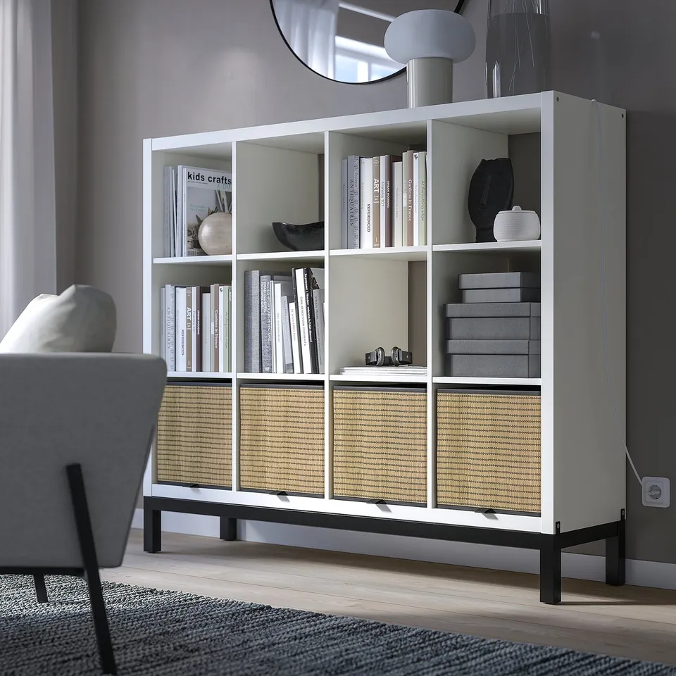 White KALLAX bookshelf with books, decor, and a basket, larger than a nearby grey chair.