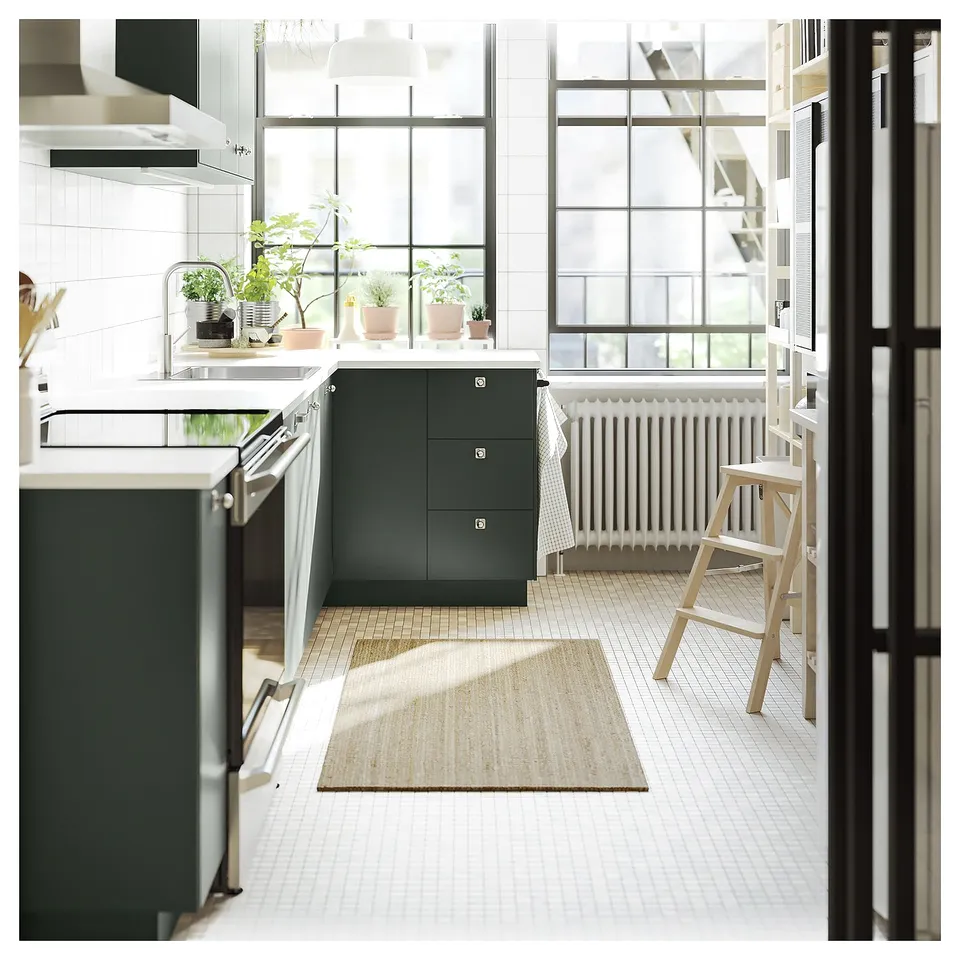 Modern kitchen with white/green cabinets, large windows, plants, stools, radiator, jute rug.