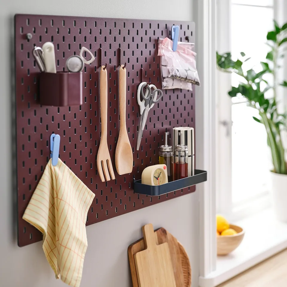 Organised kitchen tools on a dark brown SKÅDIS pegboard. Includes wooden utensils, scissors, clock, and shelf with bottles.