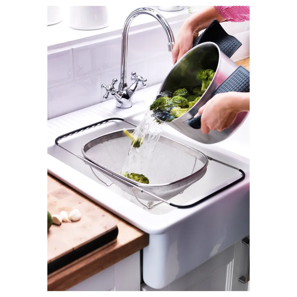 Person rinsing broccoli in sink using stainless steel colander with adjustable handles.