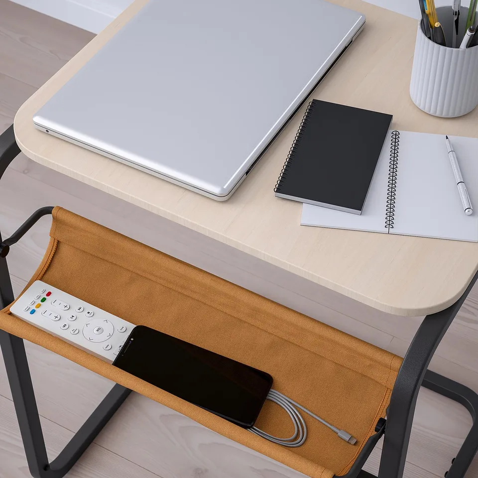 A wooden desk with a laptop, notebook, and phone. A fabric pouch hangs from the desk, holding a remote.