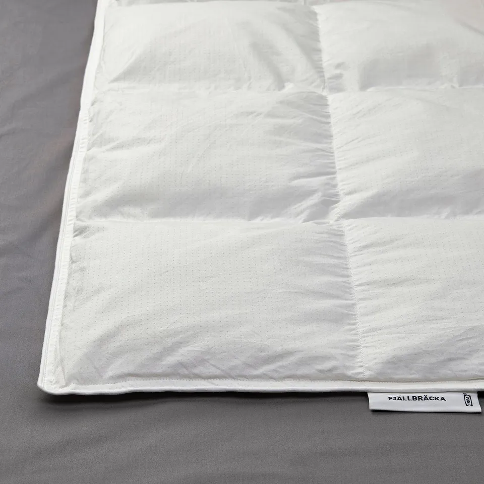 Clean white duvet neatly folded on grey bedding. Tag reads FJÄLLBRÄCKA with recycled down filling.