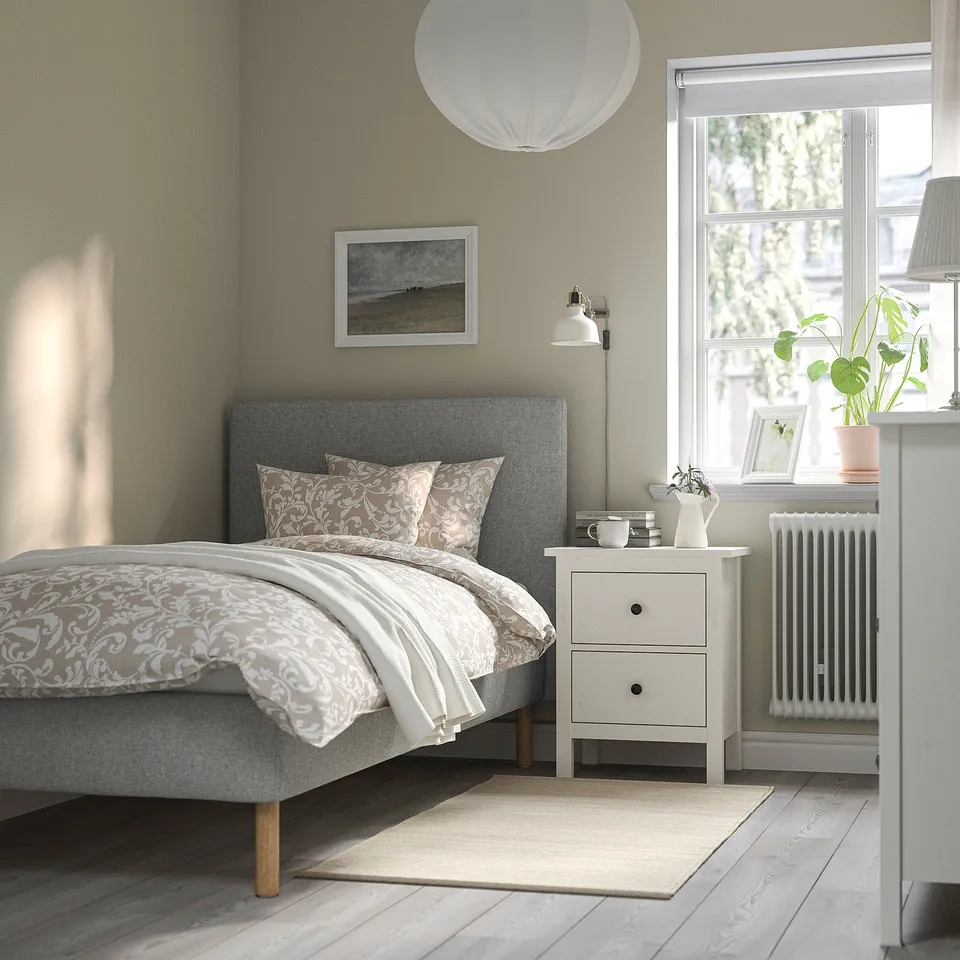 Minimalist bedroom: grey walls, floral bed, large window, bedside table with plant, rug, radiator.