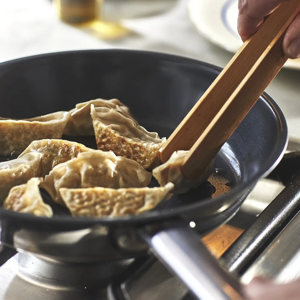 Person using wooden chopsticks to pick dumplings from a non-stick frying pan on a stove.