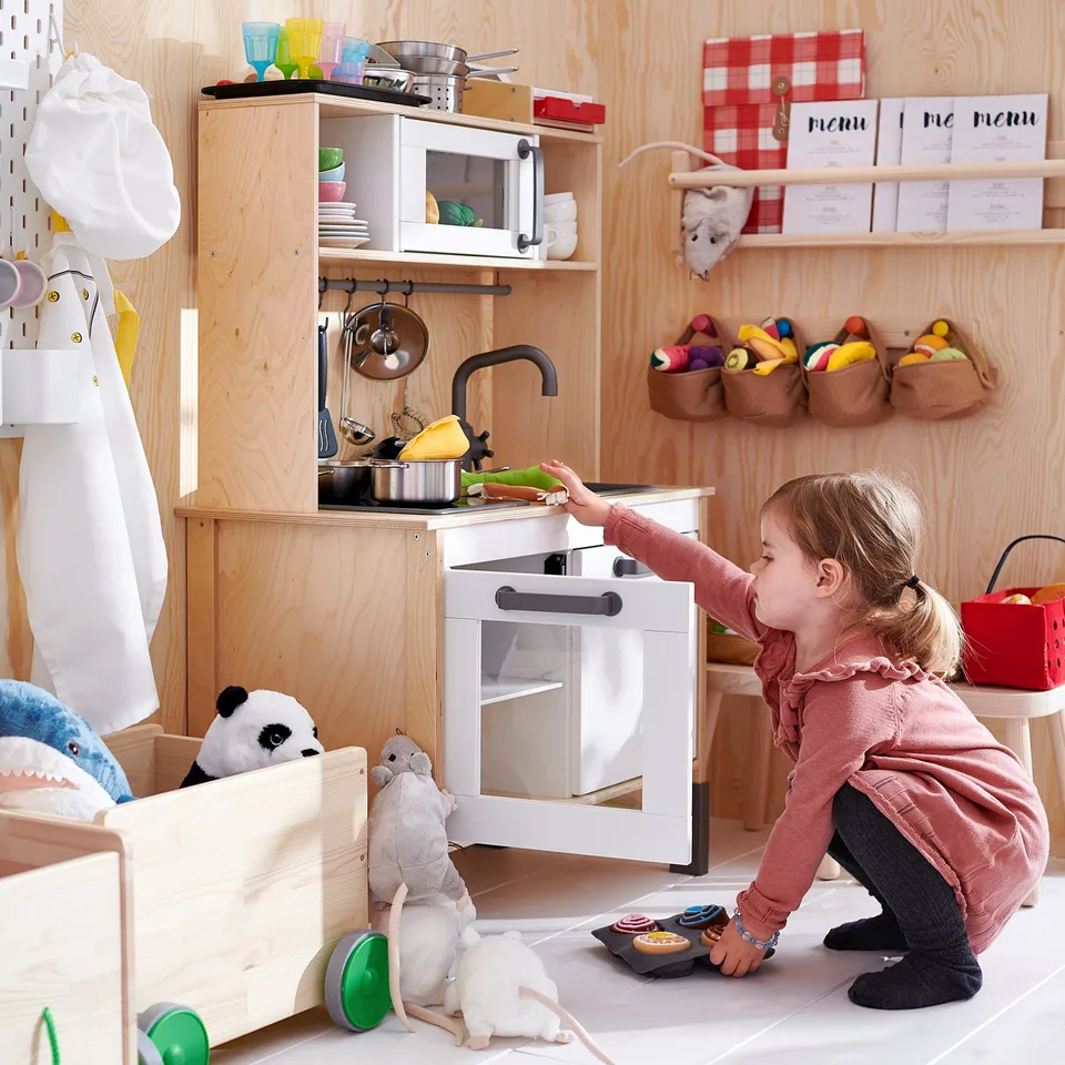Girl plays with toy stove in wooden play kitchen, surrounded by colourful toys.