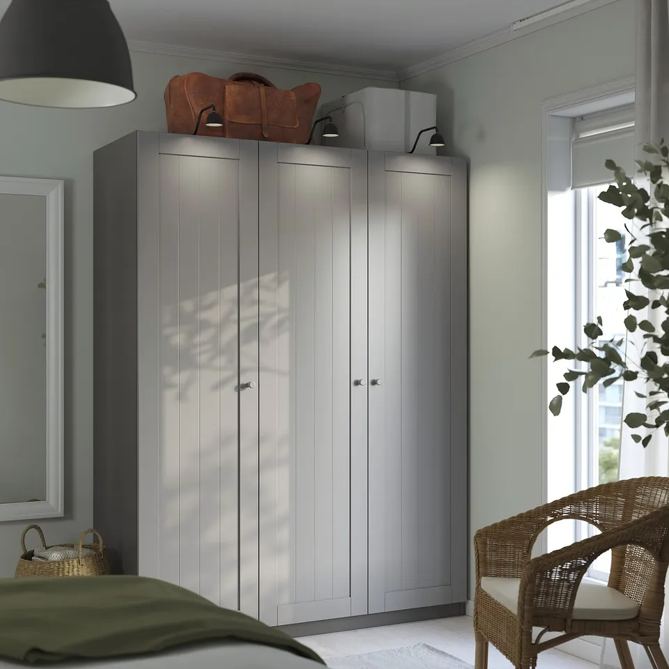 Modern grey PAX wardrobe with two doors, tall and sleek, positioned beside a window with blinds, a wicker chair, and a hanging plant.