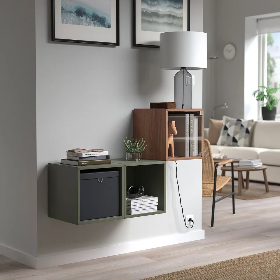 Modern living space with grey EKET storage, books, headphones, plant, paintings, and lamps.