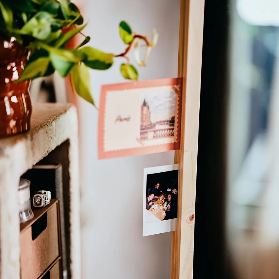 A modern mirror with photos attached. It has sleek, wooden sides and sturdy hooks. A plant sits nearby.