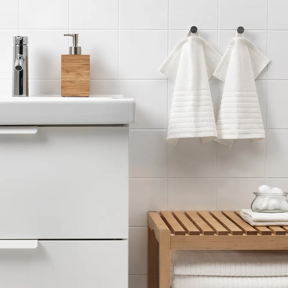 Modern bathroom with white cabinets, sink, VÅGSJÖN towels, bench, and lotion dispenser.