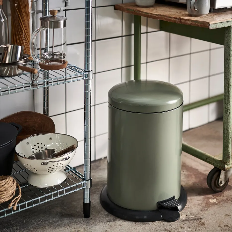 Green rubbish can in kitchen, next to metal shelf with utensils.
