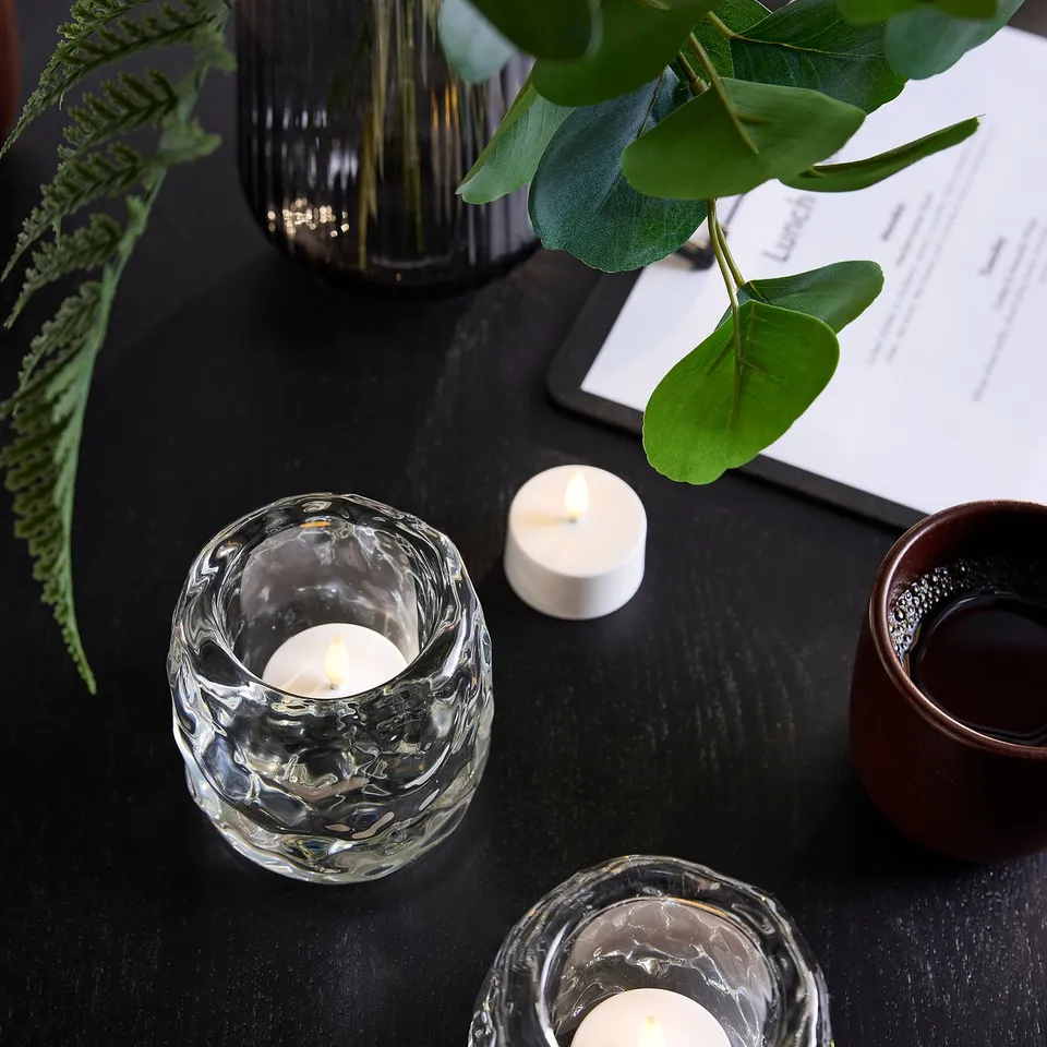 Two lit glass SKATGÅS tealights on black table, surrounded by a black lamp, brown cup, and greenery, tealights are small.