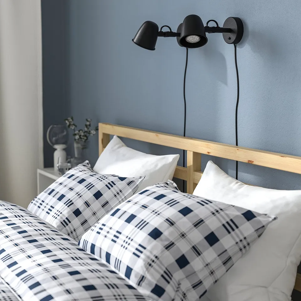 Bedroom scene with TARVA bed frame, light blue walls, plaid bedding, two black wall-mounted reading lights above bed.