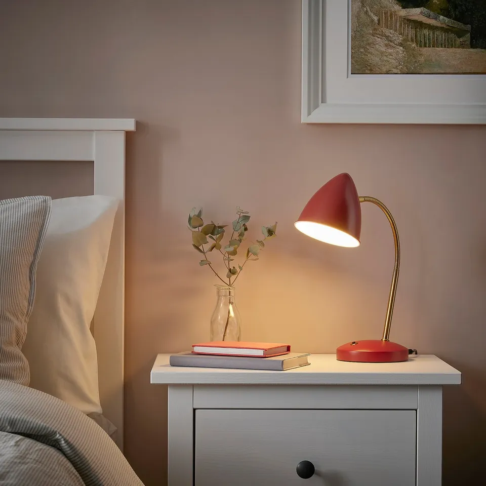 Red lamp, books, vase with dried flowers on bedside table near bed.