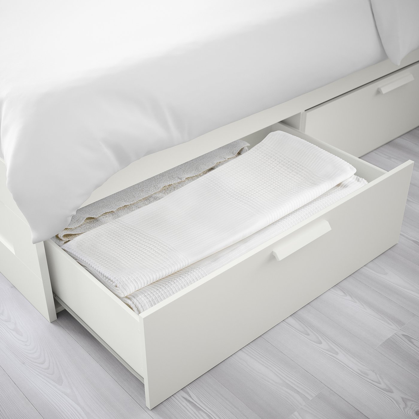 Open white bed storage drawers, showing folded white linens inside, set on light wooden floors.
