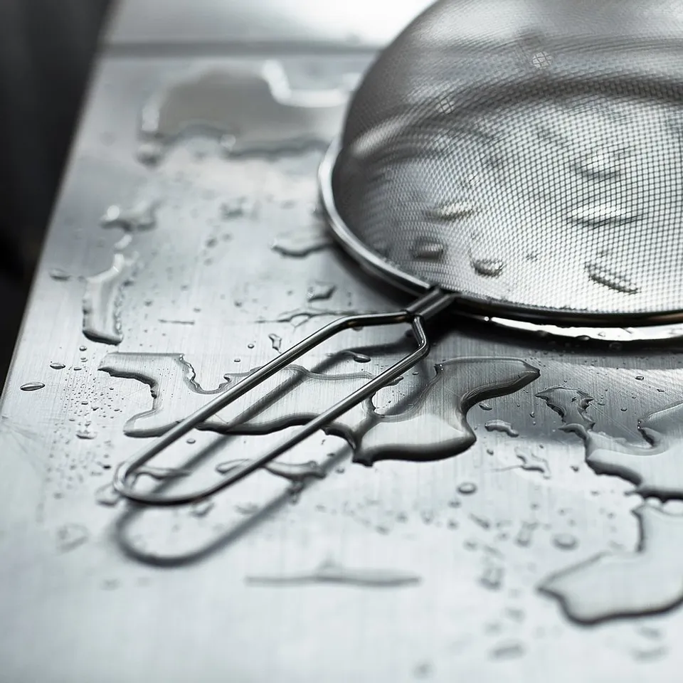 Stainless steel sifter and tongs on wet, metallic surface. Close-up, monochrome image emphasising textures and reflections.