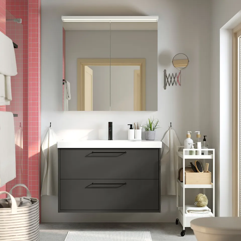 Modern grey vanity with mirror, white top, black handles, pink wall, under-sink cart, towels.