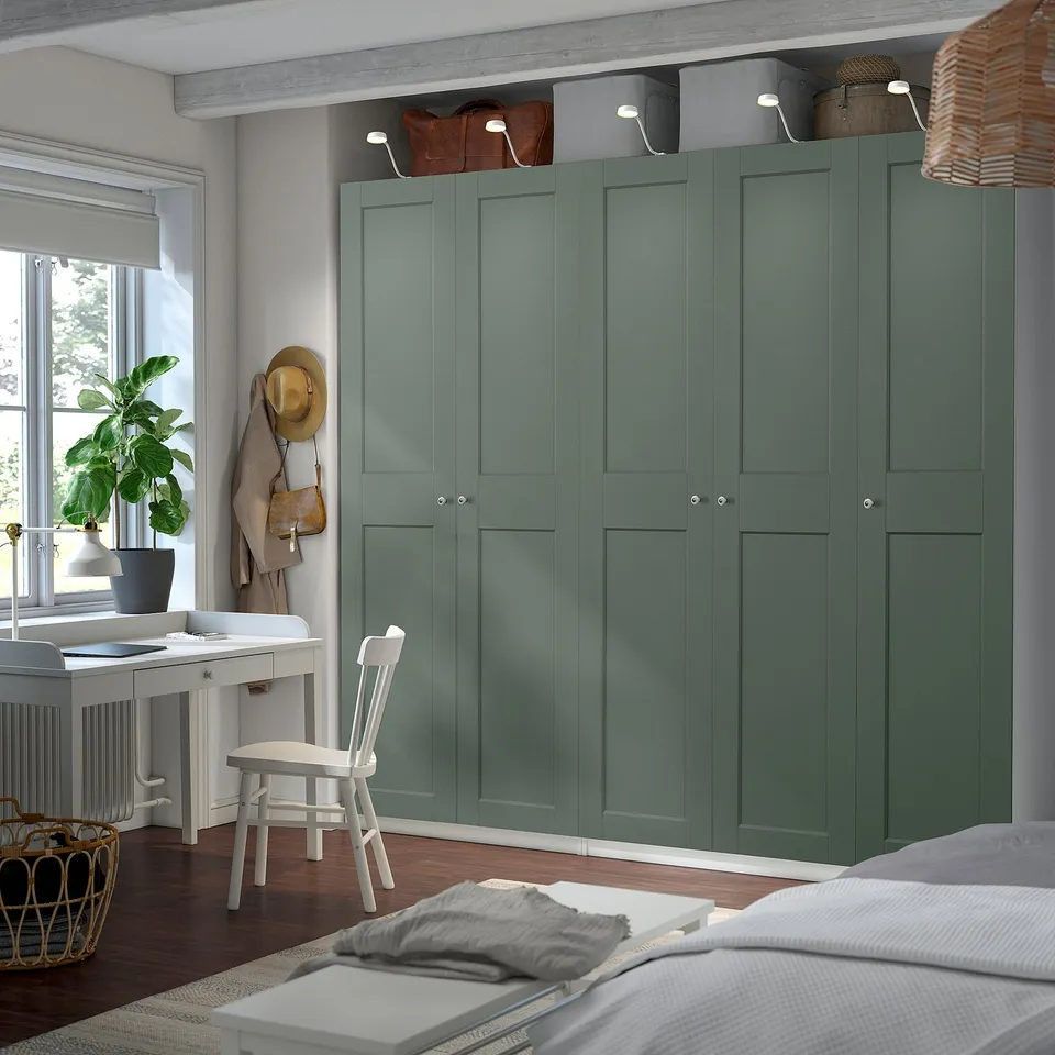 Bedroom with green PAX wardrobe, desk, and bed. Wardrobe has grey-green finish, mesh basket, and lights.