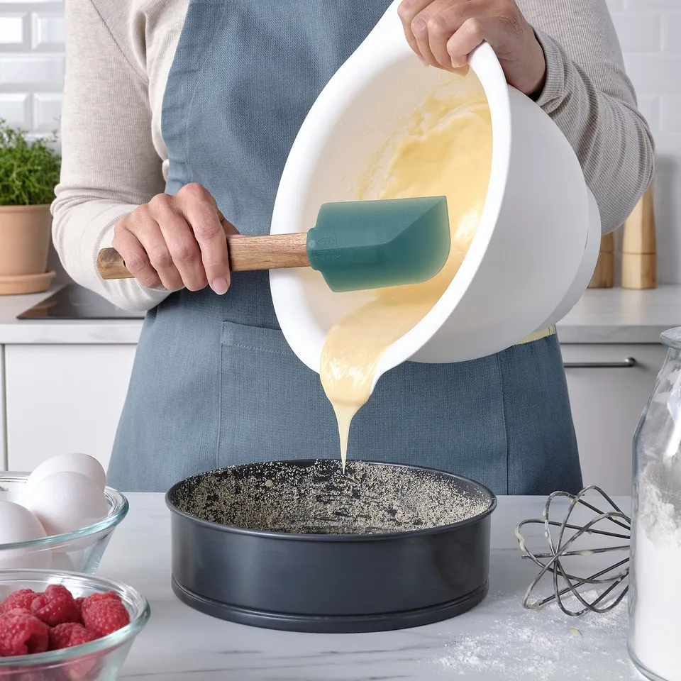 Person pours batter from a white bowl into a round pan using a green spatula. Other ingredients and kitchen tools are nearby.