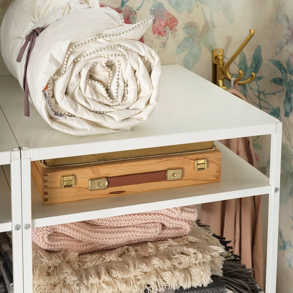 White JONAXEL storage unit with rolled white linens and folded pink blankets. Gold-accented wooden box on middle shelf.