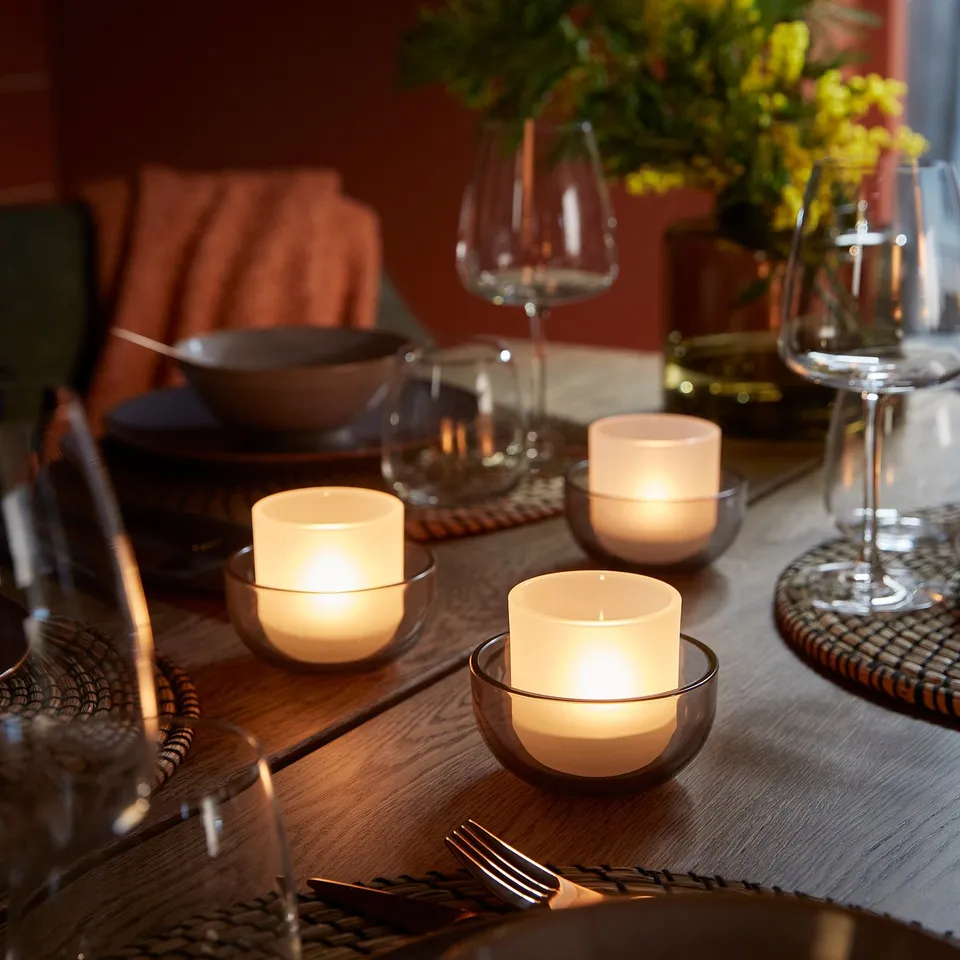 Table setting with wooden surface, lit candles in glass holders, wine glasses, plates, cutlery, and a vase with flowers.