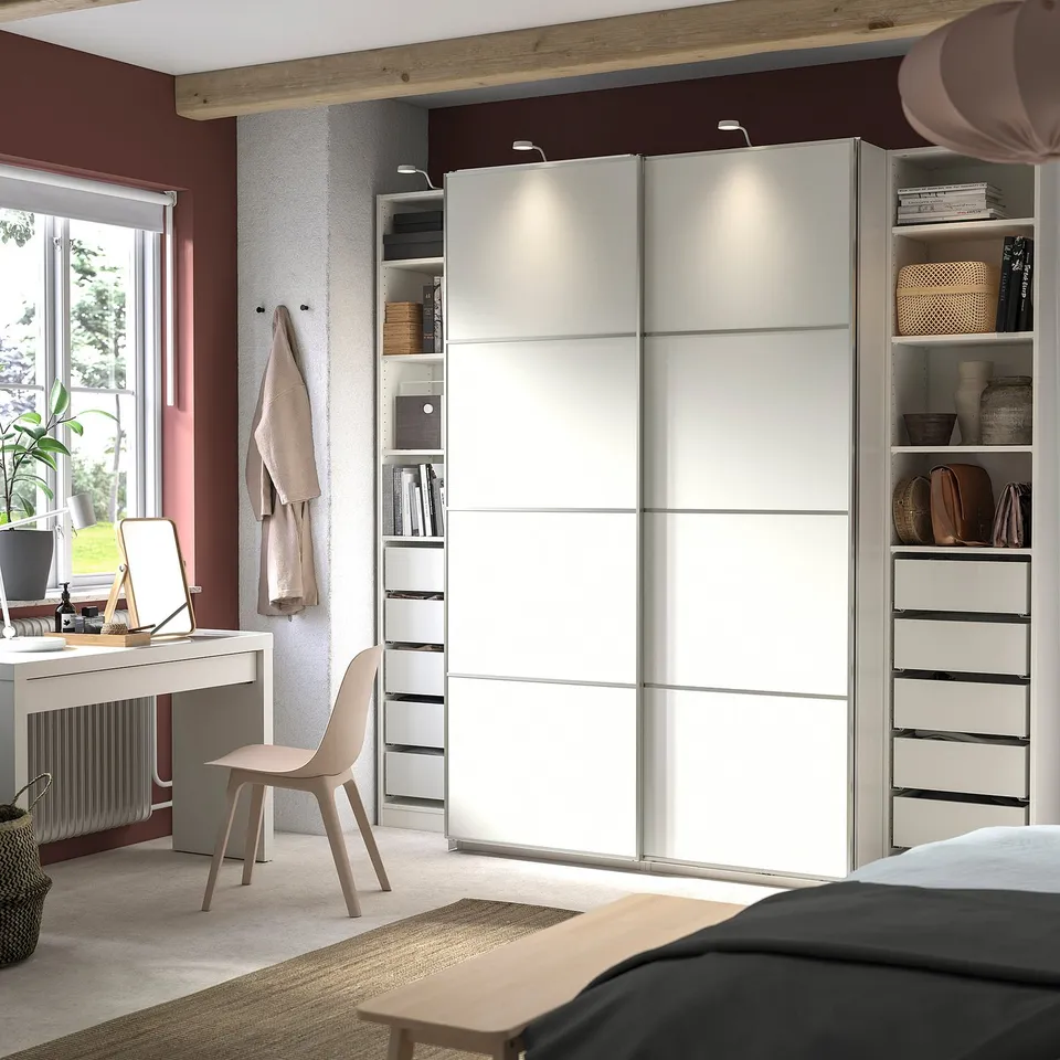 Modern bedroom with white PAX wardrobe, sliding doors, and organised shelves. Neutral decor with a desk and chair, natural light from window.