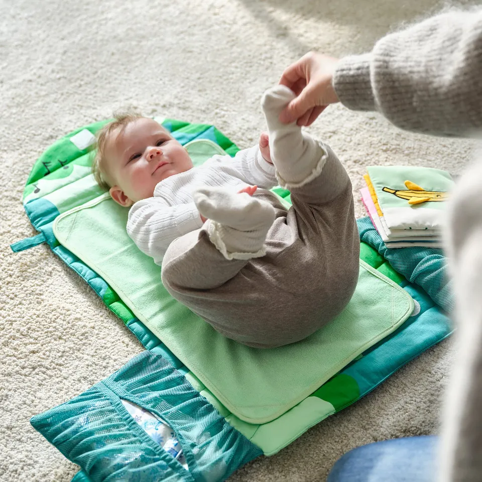A baby lies on a green changing mat. An adults hand reaches towards the baby. The mat has pockets for diapers.