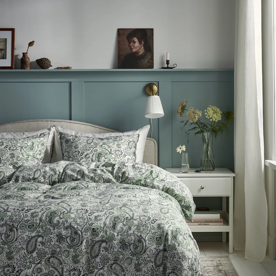 Teal bedroom with teal walls, white bedding, green paisley quilt, and headboard. Bedside table with vases and curtains.