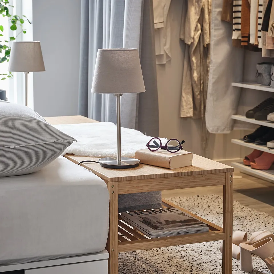 Bedside table with wooden frame and white table lamp with grey shade, beside a bed.