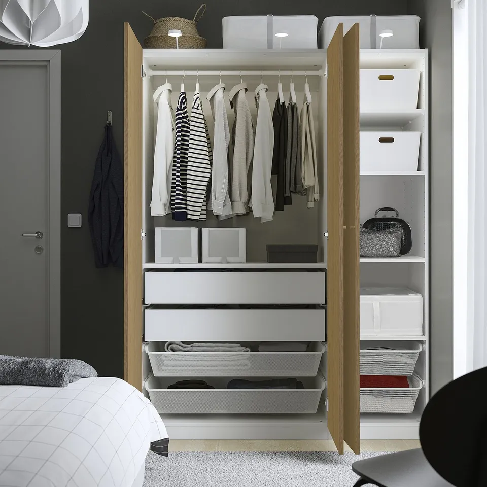 A modern, white PAX wardrobe with open doors displaying clothes, drawers, boxes, and mesh baskets for organised storage.