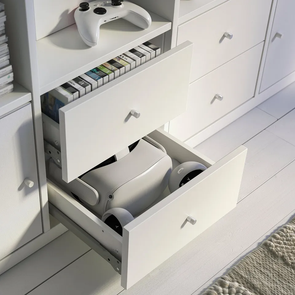 White KALLAX unit with open drawers holding gaming controllers and cds; top shelf holds a game controller.